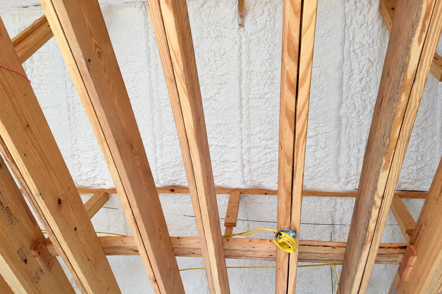 White spray foam seals the underside of a roof between exposed wooden rafters, with a small loop of yellow wire visible.