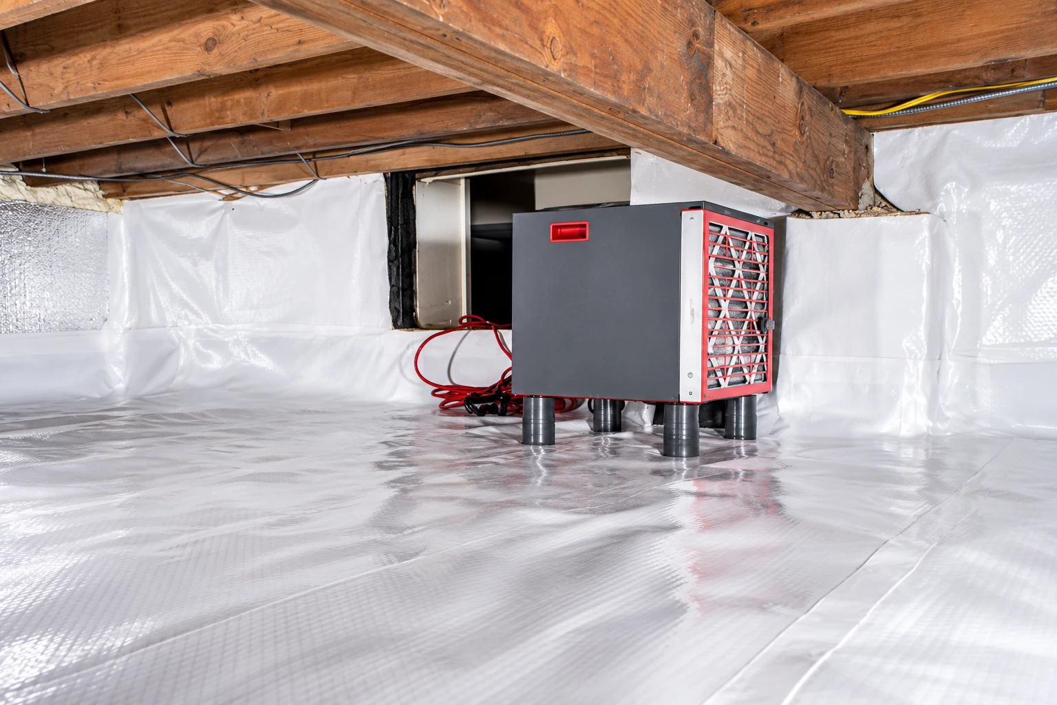Gray-and-red machine sits on a plastic-lined crawl space floor beneath exposed wooden joists.