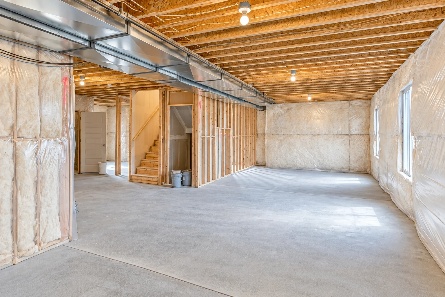 An unfinished basement has white insulation along its walls, exposed ceiling joists, metal ductwork, and a bare concrete floor.