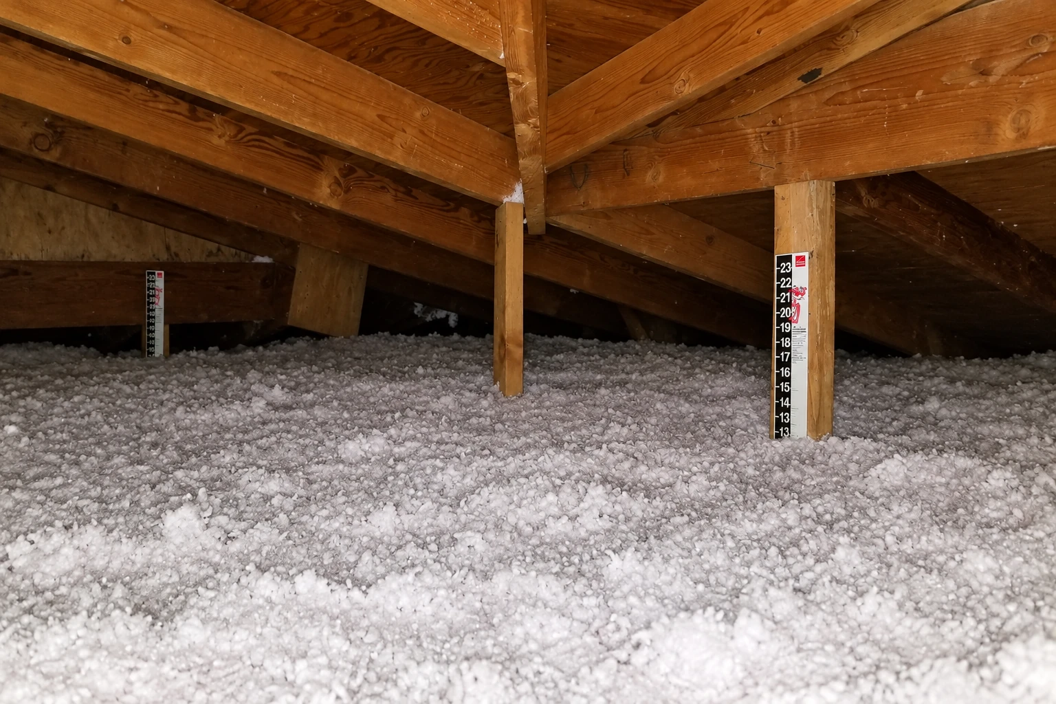 Loose white blown-in insulation covers an attic floor beneath wooden roof trusses.