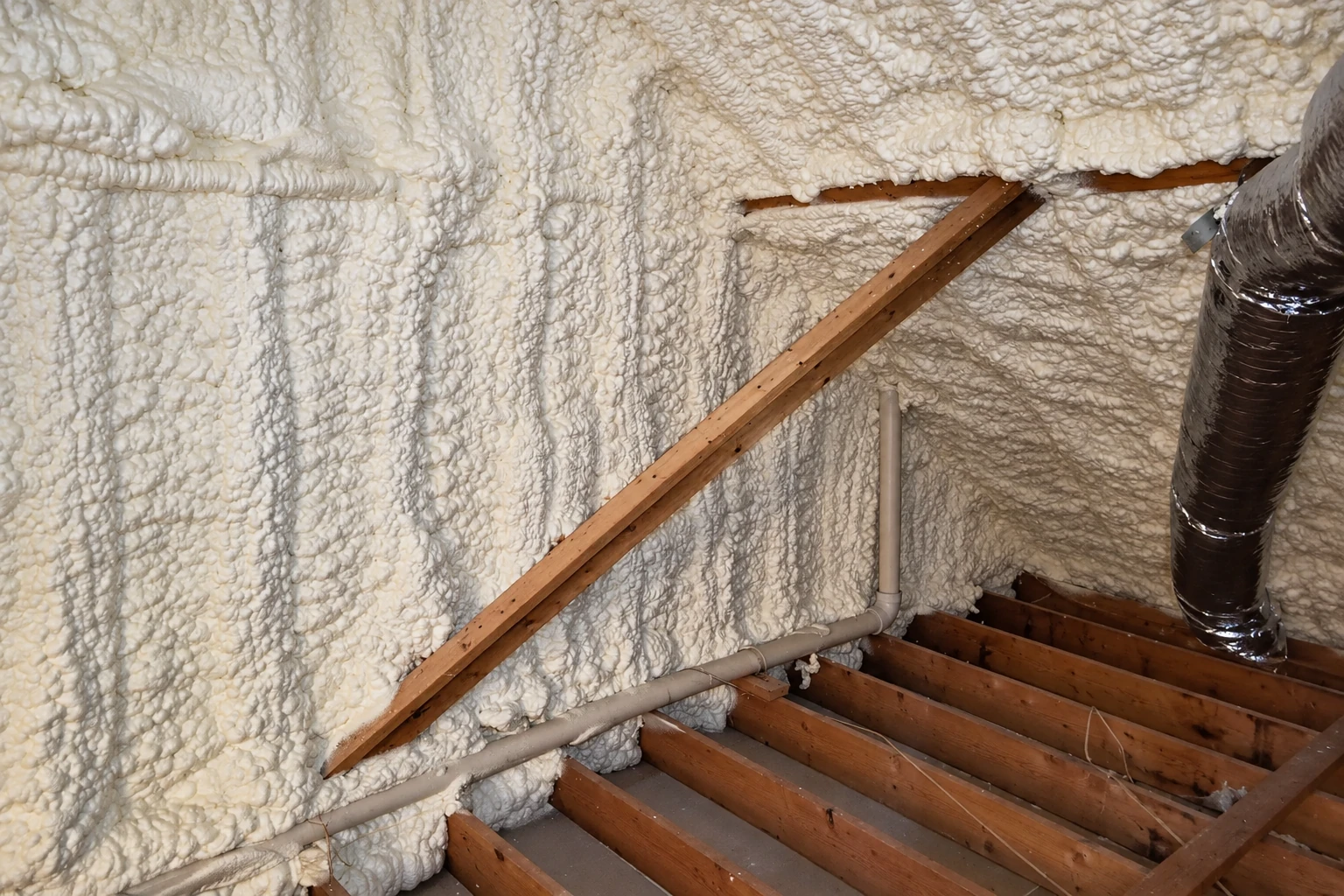 Exposed wooden framing and pipes surrounded by thick, uneven white closed-cell spray foam insulation.