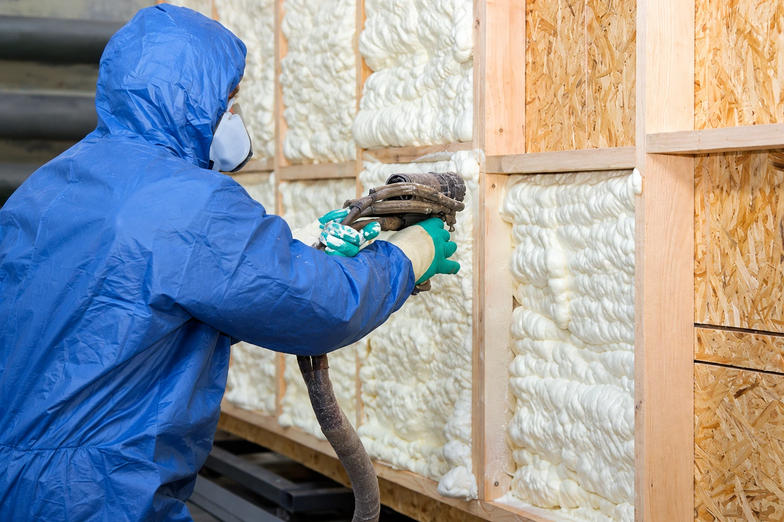 A worker in protective gear sprays foam insulation into framed wall cavities in a building under construction.