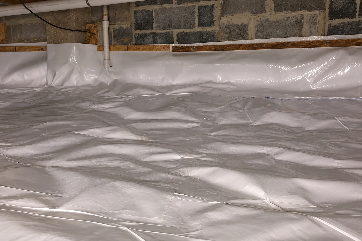 White vapor barrier covers the crawl space floor and lower masonry walls beneath a pipe.