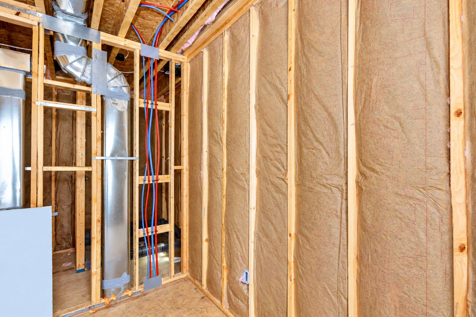 Tan batt insulation fills the wall cavities of an unfinished room beside exposed framing, ducts, and red and blue pipes.